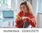 Young adult smiling pretty woman sitting on chair holding smartphone using cellphone modern technology, looking at mobile phone while remote working or learning, texting messages at home office.