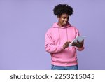 Small photo of Young happy cool curly African American teenager student boy wearing pink hoodie holding pad using digital tablet computer technology browsing, elearning standing isolated on light purple background.