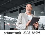 Smiling mid aged business man wearing suit standing outside office holding digital tablet. Mature businessman professional holding fintech device looking away thinking or new business ideas solutions.