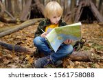 Small photo of Little boy scout is orienteering in forest. Child is sitting on fallen tree and looking on map on background of teepee hut. Concepts of adventure, scouting and hiking tourism for kids.