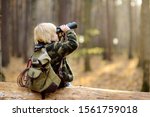 Small photo of Little boy scout with binoculars during hiking in autumn forest. Child is sitting on large fallen tree and looking through a binoculars. Concepts of adventure, scouting and hiking tourism for kids.