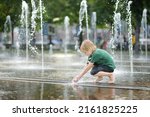 Little boy plays in the square between the water jets in the city fountain at sunny summer day. Active summer leisure for kids in a big city.