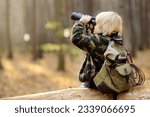 Small photo of Little boy scout with binoculars during hiking in autumn forest. Child is sitting on large fallen tree and looking through a binoculars. Concepts of adventure, scouting and hiking tourism for kids.