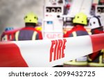 Small photo of Scene of incident is cordoned off with red and white tape, reading ‘fire’. Out of focus in the background are three firefighters, two fire engine appliances and smoke billowing through the air.