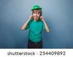 Small photo of European appearance teenager boy in T-shirt with green hat stretches out his tongue and ears on gray background, foolery, teasing