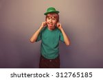 Small photo of European appearance teenager boy in T-shirt with green hat stretches out his tongue and ears on gray background, foolery, teasing retro
