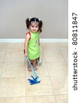 Small photo of Adorable little girl pushes her mop back and forth cleaning the floors. She is learning household chores from her mom. She is wearing a green apron and holding a blue, play, mop.