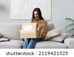 Happy young woman sit on couch in living room unpack cardboard box buying goods on Internet, smiling excited millennial girl open carton parcel order, shopping online, good delivery concept