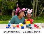 Small photo of Cheerful african american girl and grandmother wearing bunny ears while painting eggs on easter day. unaltered, lifestyle, easter day, art, celebration, family, cultures and holiday concept.