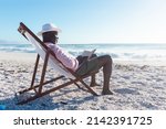 Small photo of Side view of african american senior man reading book sitting on folding chair at beach. unaltered, hobbies, active lifestyle, enjoyment and holiday concept.