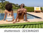 Small photo of Rear view of a couple sitting in the garden on the side of a swimming pool wearing beachwear and sunbathing