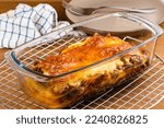 High angle view of freshly baked homemade lasagna in transparent tray on cooling rack with brown ceramic dish and metal fork on wooden table.
