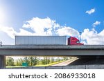 Industrial powerful red big rig classic semi truck transporting commercial cargo in dry van semi trailer running on the concrete bridge across the wide highway road on the cloud sky background