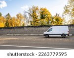 Commercial compact cargo small size white mini van delivering cargo to client running on the local road with autumn yellow trees and concrete protection fence on the side