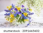 Small photo of A bouquet of spring blue, yellow flowers in a vase on the table. Pansies, forget-me-nots, primroses, bird cherry, violets, muscari. Postcard, blur, selective focus.