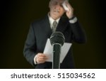 Small photo of Microphone in sharp focus with a nervous sweaty public speaker or politician blurred in the background preparing to make a speech as he wipes his brow