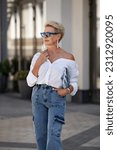 Small photo of Beautiful mature woman with chic blonde bob outdoors in city street wearing trendy white shirt, cargo jeans pants and fashionable accessories, stylish glasses and clutch. Fashion style portrait.
