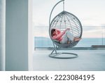 Beautiful middle-aged barefoot woman lying in comfortable hanging chair on open house terrace and enjoying the sunset sun rays. Modern home lifestyle and mental health concept image