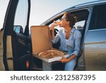 Midle-aged woman eating just cooked italian pizza sitting on driver car seat during meal break and enjoying sunset light. Auto traveling, fast food eating or car jorney lunch break concept image.