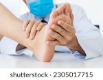 Small photo of Examination of a young woman by an orthopedist.White uniform male doctor holding a girl's foot in his hands. Flat feet, injury. Foot treatment. Pain from uncomfortable shoes.Medical orthopedic unit.