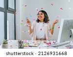Small photo of Young business woman having fun time catching confetti sitting at the desk in the office. Party time on the work place. Selective focus.