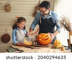Smiling father showing his little daughter how to carve pumpkin with spooky face, dad with child creating this iconic symbol of Halloween holiday jack-o-lantern while standing behind table in kitchen