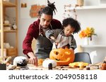 Cute happy little african american boy carving Halloween pumpkin with father at home, mixed-race family son and dad making Jack-o-Lantern together while preparing decorations for Saints Day party