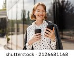 Young positive businesswoman in glasses with take away coffee cup in hand using mobile phone while standing on sunny street near office building, chatting online with friends during break at work