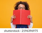 Small photo of Mischievous ethnic curly boy in glasses hugging textbooks and looks out from behind the book during school studies against yellow background