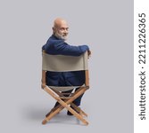 Small photo of Confident film director sitting on the director's chair and looking at camera: video production, filmmaking and film industry concept
