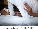 Small photo of Handsome young male insomniac covering his head and ears trying to block out the sound with a pillow as he is kept awake by loud noises around him