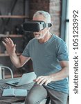 Small photo of New experience. Concentrated senior unshaken man sitting in the empty room on the table tasting VR glasses and shaking hands.