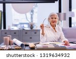 Small photo of CEO in office. Prepossessing sitting grey-haired business lady in years wearing white shirt and massive earrings and smilingly holding glasses in hand.