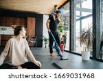 Small photo of Mother sits at easy simplified lotus pose, looking at the father vacuum cleaning apartment floor with their infant baby riding on his neck