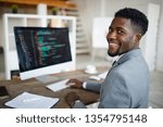 Small photo of Happy young African-american software developer in formalwear sitting by workplace in front of computer screen with decoded data