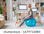 Blond mature female in activewear sitting on fitball by desk in front of computer monitor while going to exercise at home
