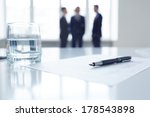 Image of business document, pen and glass of water at workplace with group of colleagues on background 