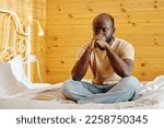 Small photo of Young tense man in jeans and t-shirt keeping clenched hands by his face while sitting on bed with his legs crossed and thinking