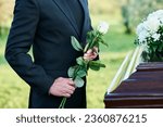 Small photo of Cropped shot of mature widower in black suit holding fresh white roses while standing by coffin with dead body of his wife at funeral