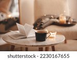 Small photo of Cup of tea with paper open book and burning scented candles on marble table over cozy chair and glowing lights in bedroom closeup. Winter holiday season.