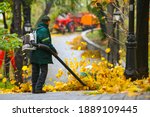 Seasonal work by city utilities in the park. A worker with a motorized backpack blower blows fallen leaves off a park path.