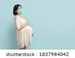 Happy Smiling Asian Pregnant woman wearing protective face mask standing over blue isolated background. Confident Young Expectant Female holding and touching her belly with copy space.