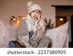 Small photo of Frozen. Sad woman sit on couch at freezing cooled house in warm cap and blanket shiver tremble with cold. Unhappy middle aged with phone lady spend time at home bad suffer of heating system problems