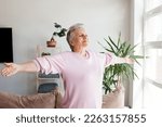 Excited elderly mature retired woman dancing in living room with widely opened outstretched arms, enjoying freedom. Overjoyed happy older female pensioner satisfied with leisure weekend time at home