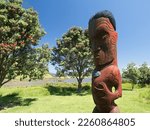 Small photo of Tawharanui- New Zealand-December 30,2022- A wooden traditional Maori carving of a heady and body in the regional park with Pohutukawa trees in background.Blue sky summer day