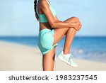 Small photo of Fitness stretches woman runner stretching glute muscle with dynamic high knee pull stretch. Athlete getting ready to run doing leg muscles warm-up standing exercise on beach pre-workout. Body closeup.