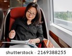 Small photo of Train commute mature Asian businesswoman working on business trip commuting to work. Happy woman sitting by window in luxury first class car reading papers