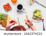 Morning habits of successful people. Day planning and healthy meal. Woman eating carrot and writing in notebook on the served for breakfast white wooden table. Top view, Selective focus
