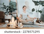 Small photo of Focus on burning candles and white Buddha statuette on tray with background of woman practicing meditation at home with cat in modern Scandi interior. Zen Composition for yoga practice, relaxation.