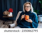 Small photo of Middle aged woman relaxing with pumpkin shaped cup of hot drink in scandy style hygge interior home with fall mood decor. Lady dreaming, enjoy calm mood without stress, well being alone. Cozy autumn